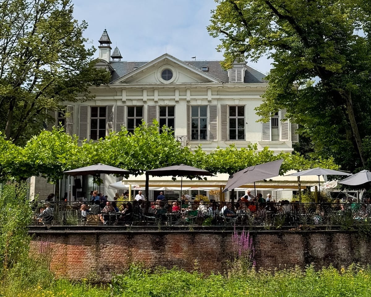A grand white estate at Middelheim Museum with outdoor seating shaded by umbrellas and greenery, bustling with cafe patrons on a sunny day.