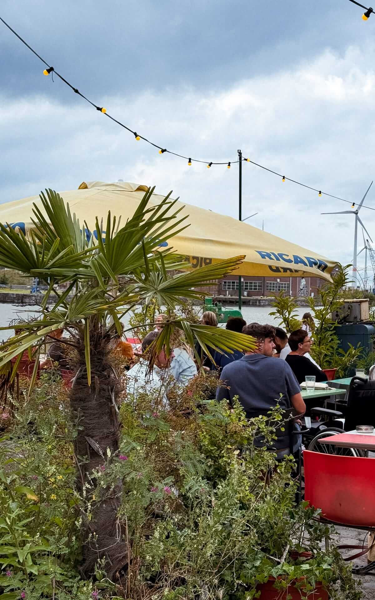 People sit under a large yellow Ricard umbrella at Paniek, a lush riverside bar in an industrial area of Antwerp.