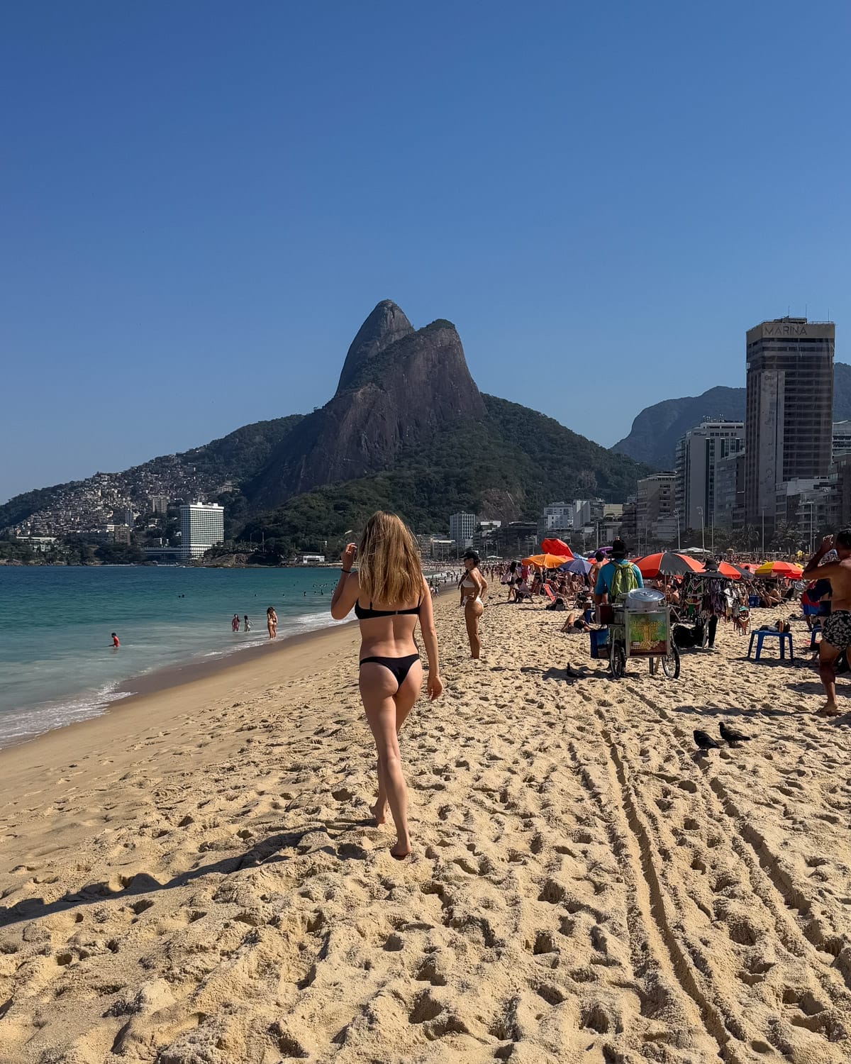 Cec walking along Leblon Beach on a sunny day with Dois Irmãos Mountain rising in the background and beachgoers under colourful umbrellas.