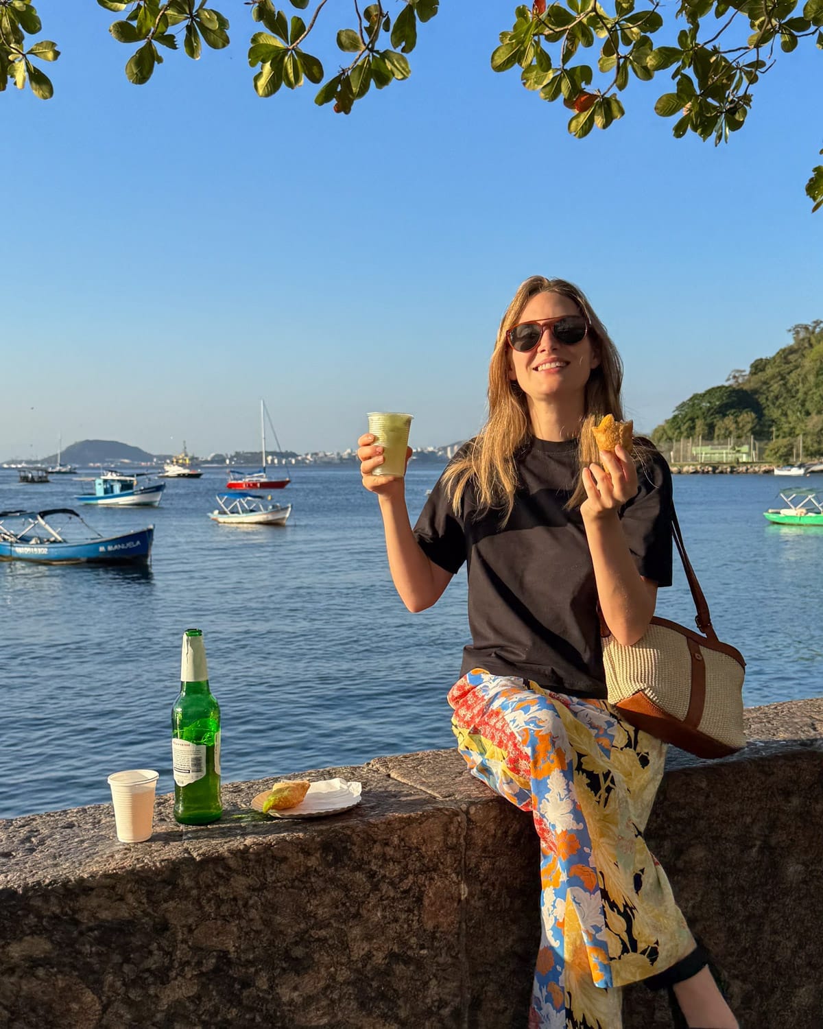 Cec sitting on the seawall enjoying cold drinks and pastéis by the waterfront at Bar Urca with fishing boats drifting by.
