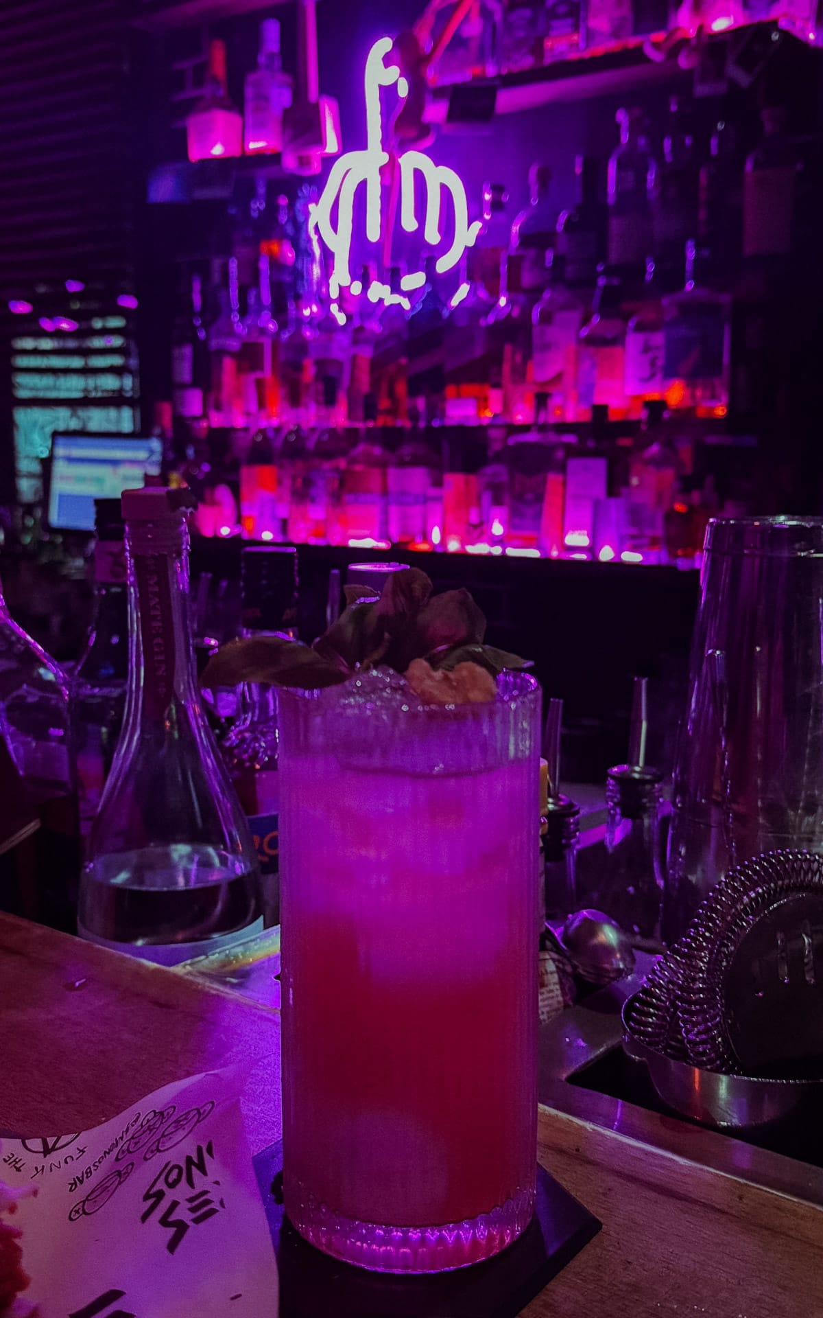 Tall pink cocktail on a bar counter with neon lights and a glowing purple sign of the middle finger in the background at Tres Monos.