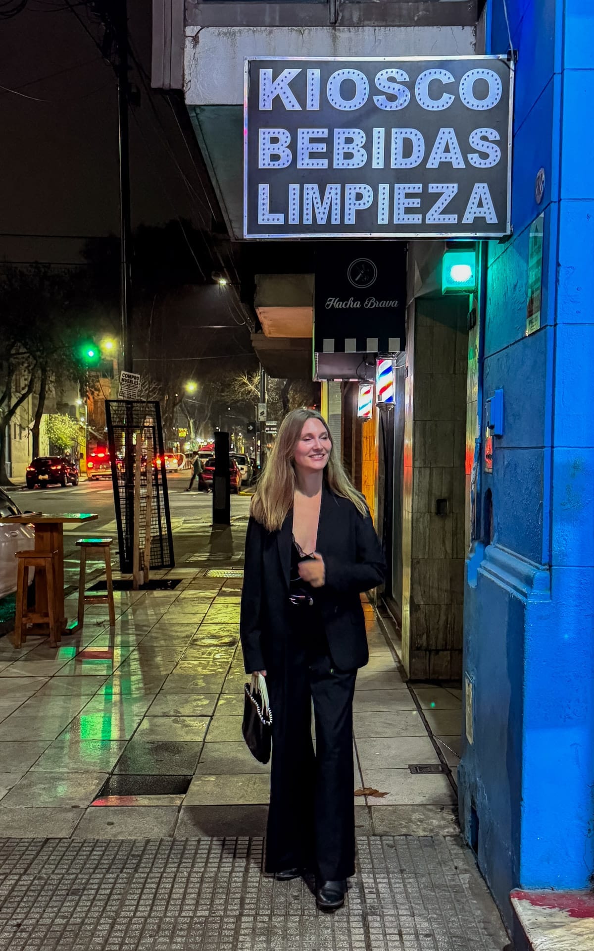 Cec dressed in black walking along a neon-lit street in Buenos Aires at night, with a kiosk sign glowing overhead.