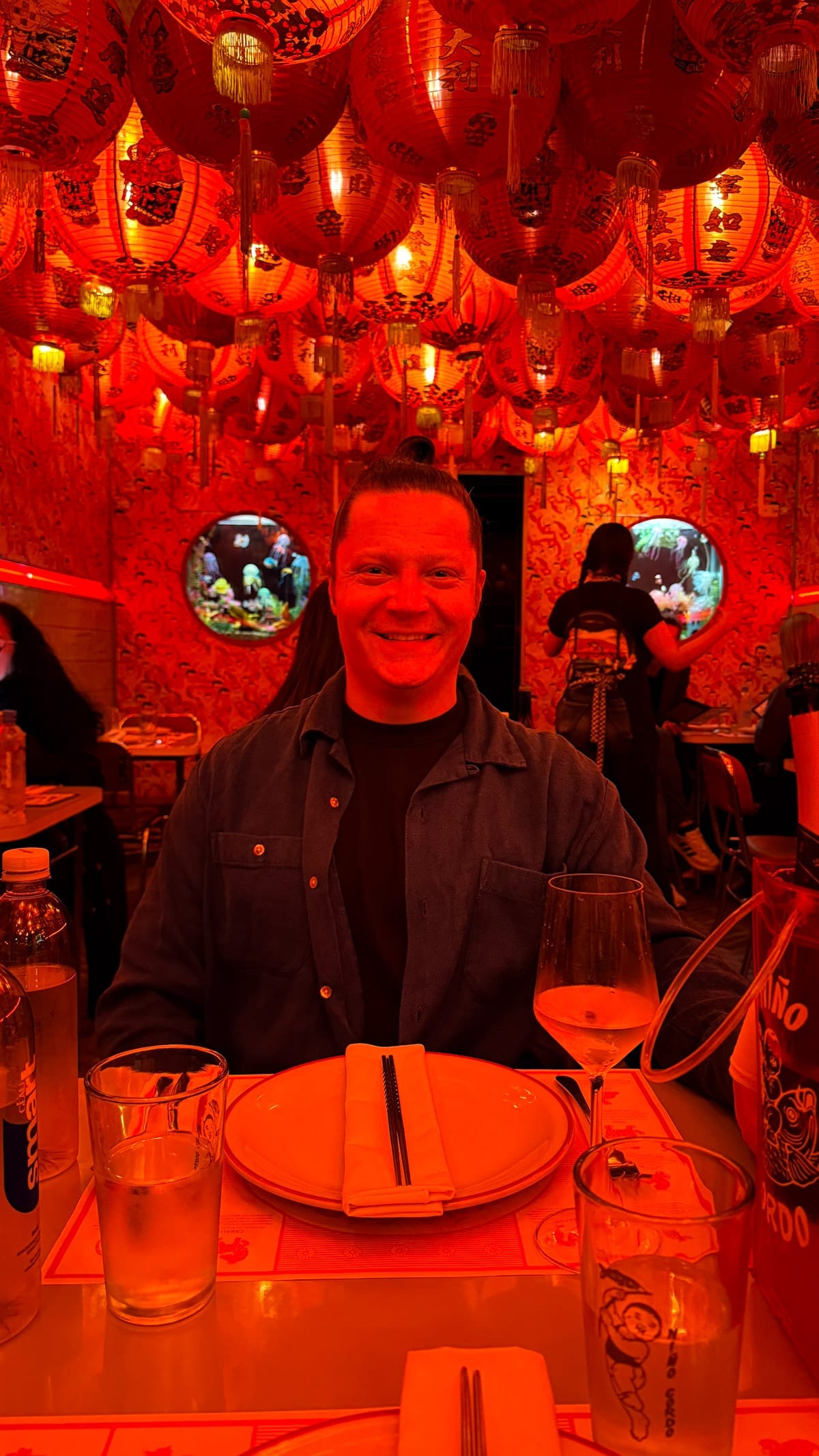 Ari smiling at a table under a ceiling of red lanterns in Nino Gordo, with aquarium windows in the background.