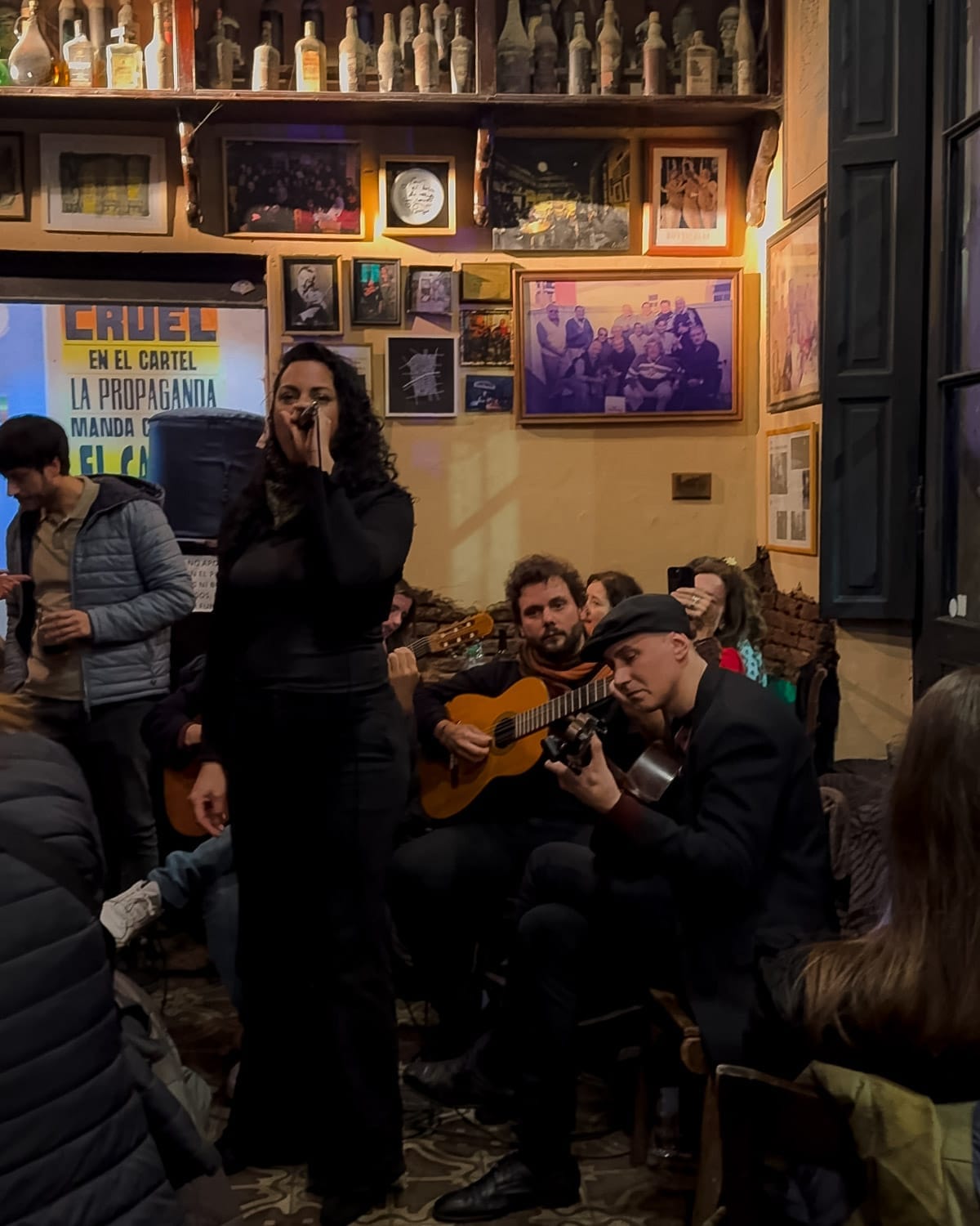 A woman sings with guitarists accompanying her at El Boliche de Roberto with vintage photos and bottles on display.