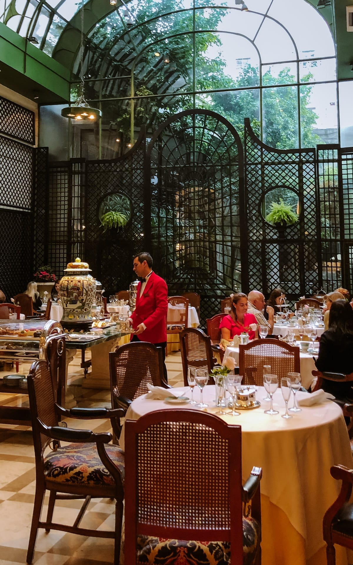 Elegant glass atrium with guests dining on white-clothed tables as a server in red offers tea service at L’Orangerie.