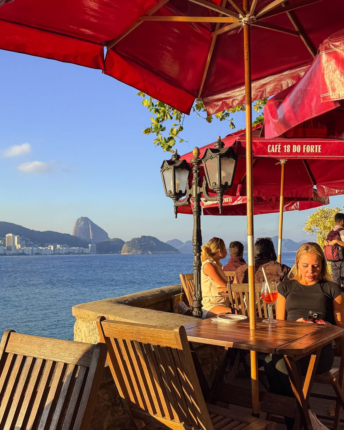 Diners enjoying drinks under bright red umbrellas at Café 18 do Forte, with sweeping ocean views and Sugarloaf Mountain rising in the distance.