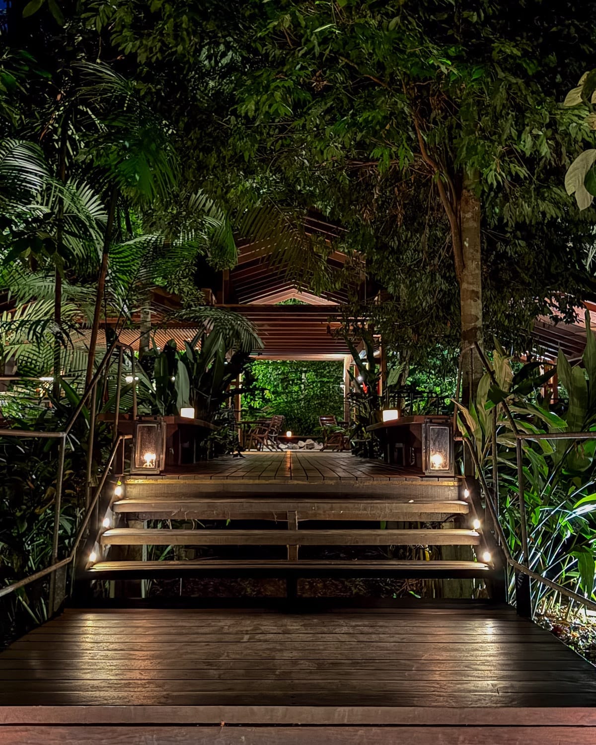 Lantern-lit wooden walkway leading into Cristalino Lodge surrounded by lush greenery at night.