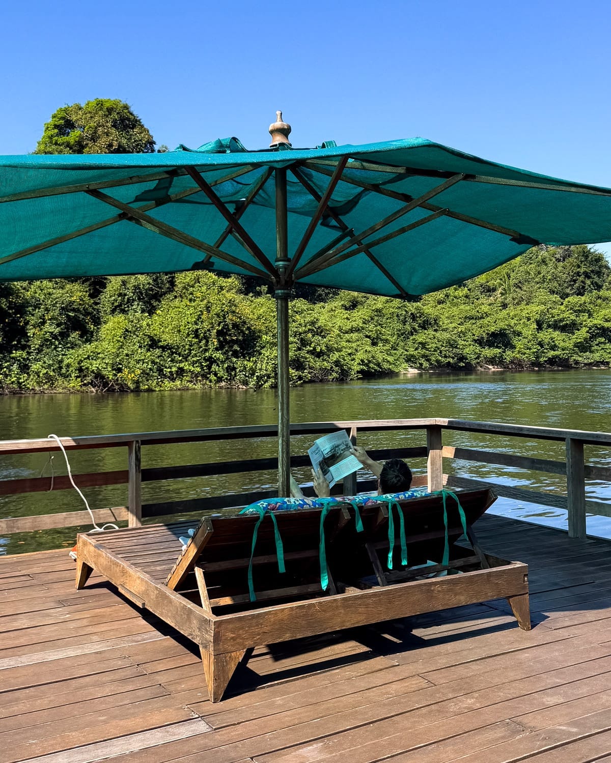 Ari relaxing on a riverside deck with lounge chairs and a large umbrella beside a jungle-lined river in the Amazon.
