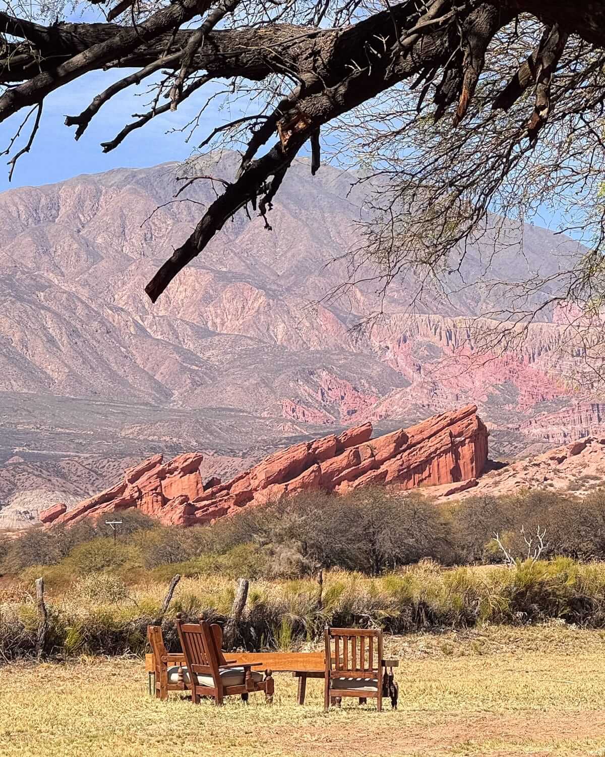 Wooden chairs and a table sit on dry grass facing layered red rock formations and distant desert mountains near Cafayate..