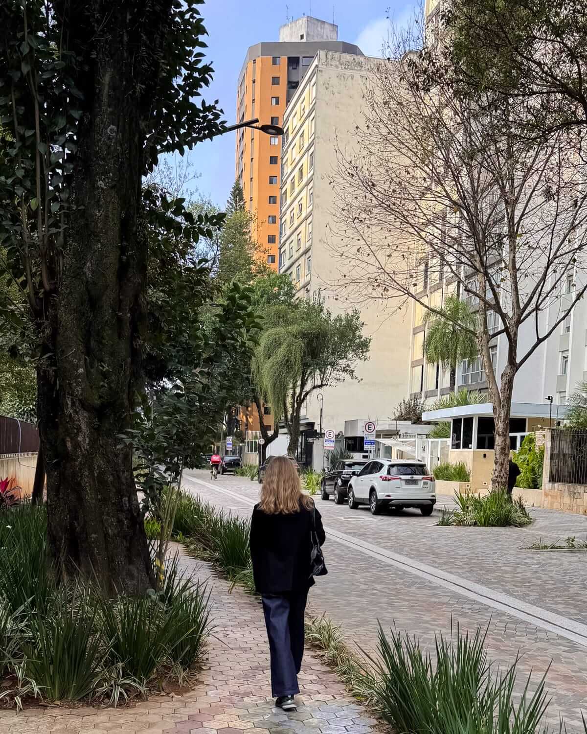 Cec walking along a tree-lined sidewalk in a quiet São Paulo neighbourhood.