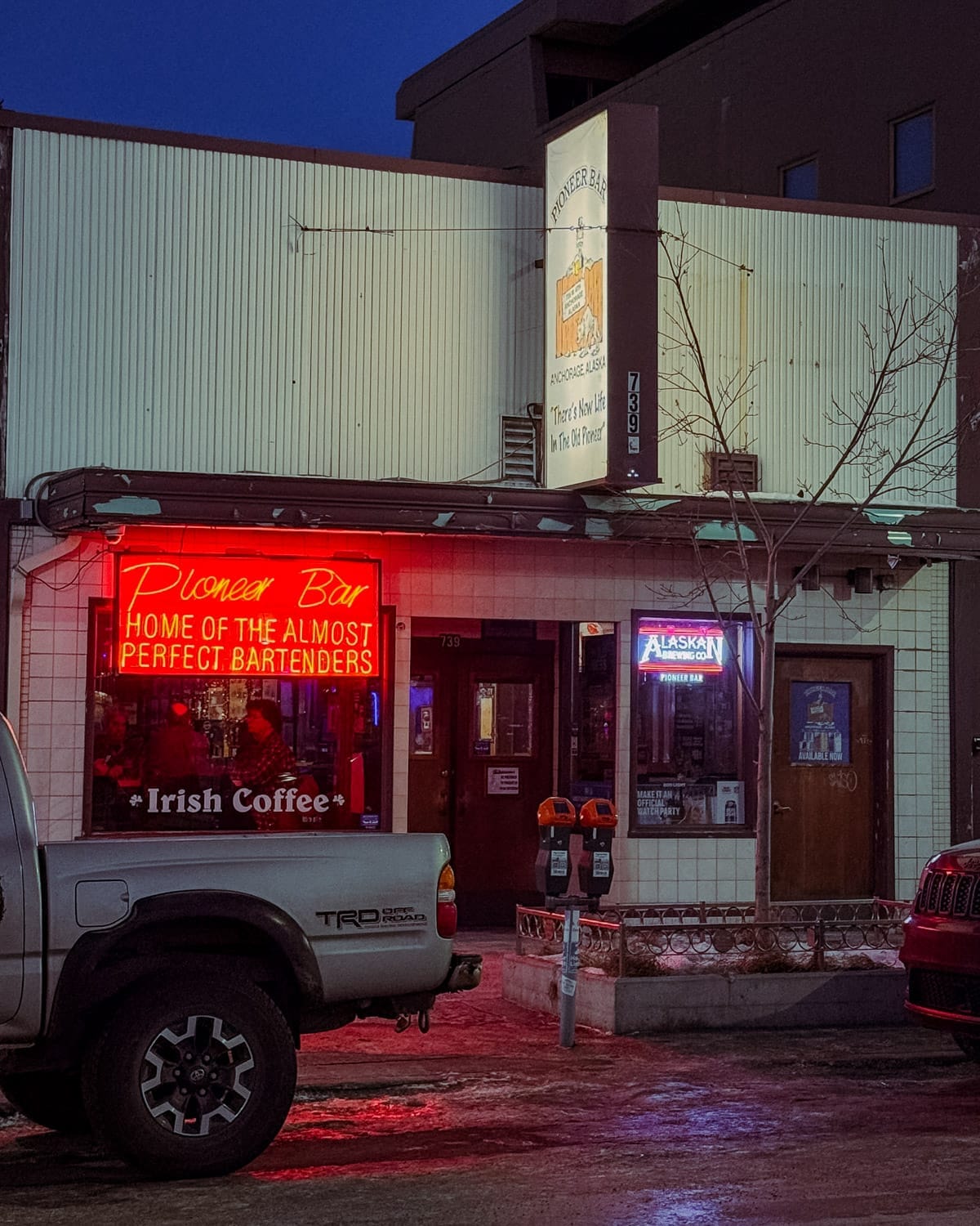 Pioneer Bar exterior in downtown Anchorage at dusk, glowing with red neon signage reading “Home of the Almost Perfect Bartenders.”