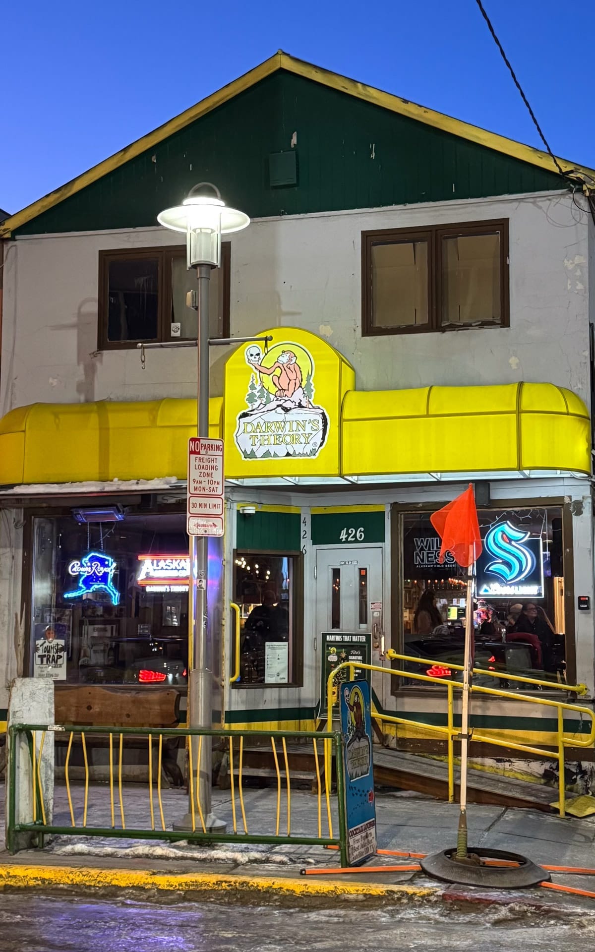 Darwin’s Theory dive bar in Anchorage with bright yellow awning and neon beer signs at night.