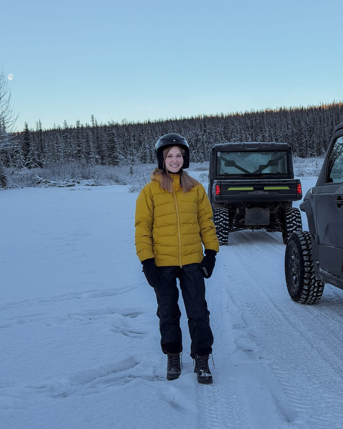 Cec in yellow winter jacket and helmet standing on a frozen river beside ATV vehicles in Alaska.