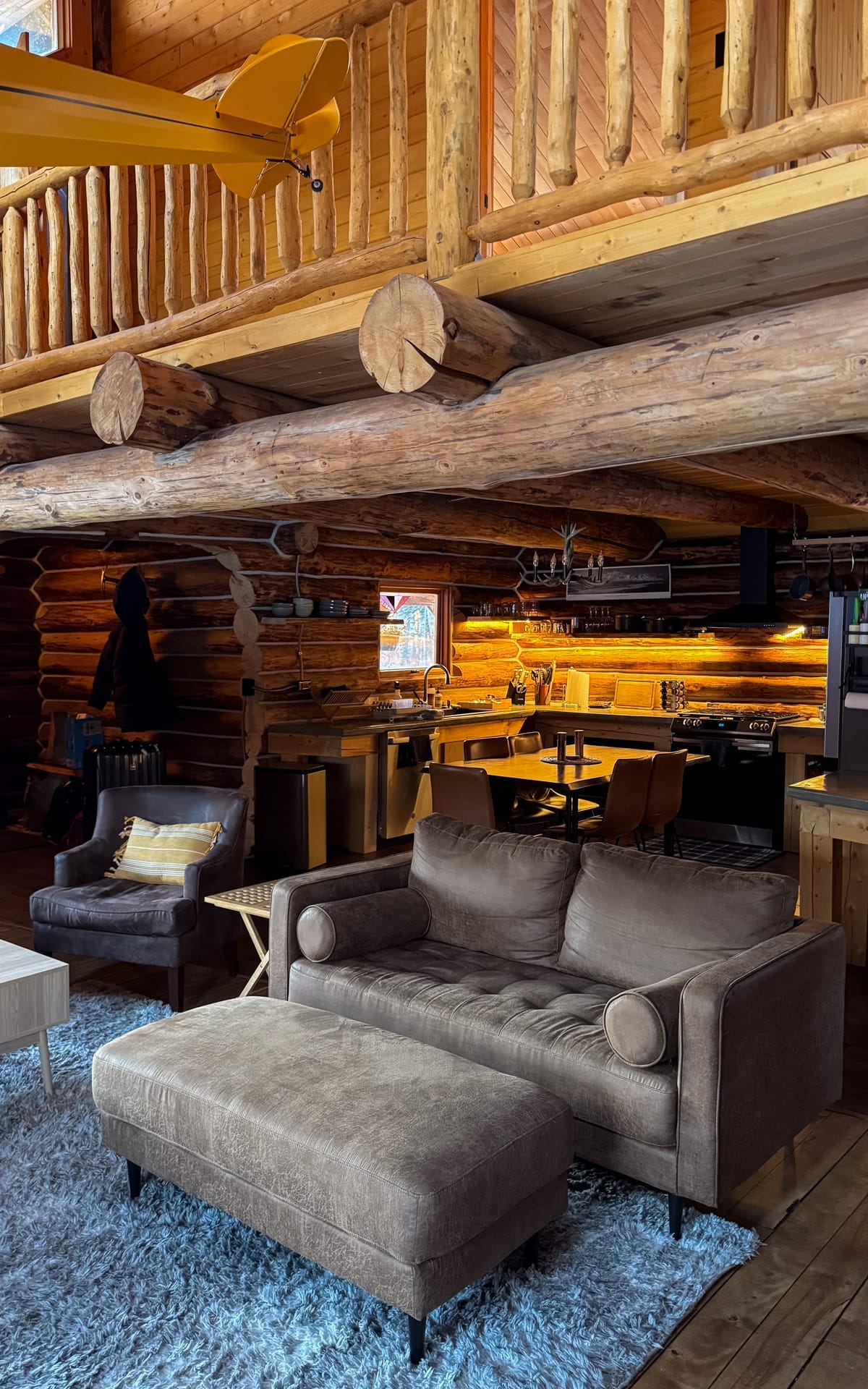 Interior of a custom log cabin in Talkeetna with leather sofa, open kitchen, exposed beams, and warm lighting.