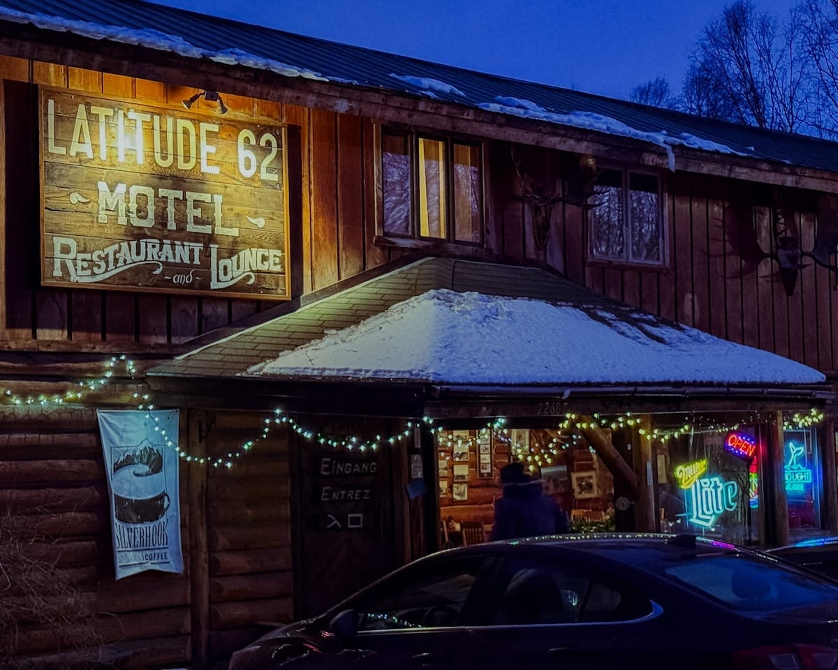Latitude 62 Motel and Restaurant exterior at dusk with string lights, snow on the roof, and glowing neon beer signs in the windows.