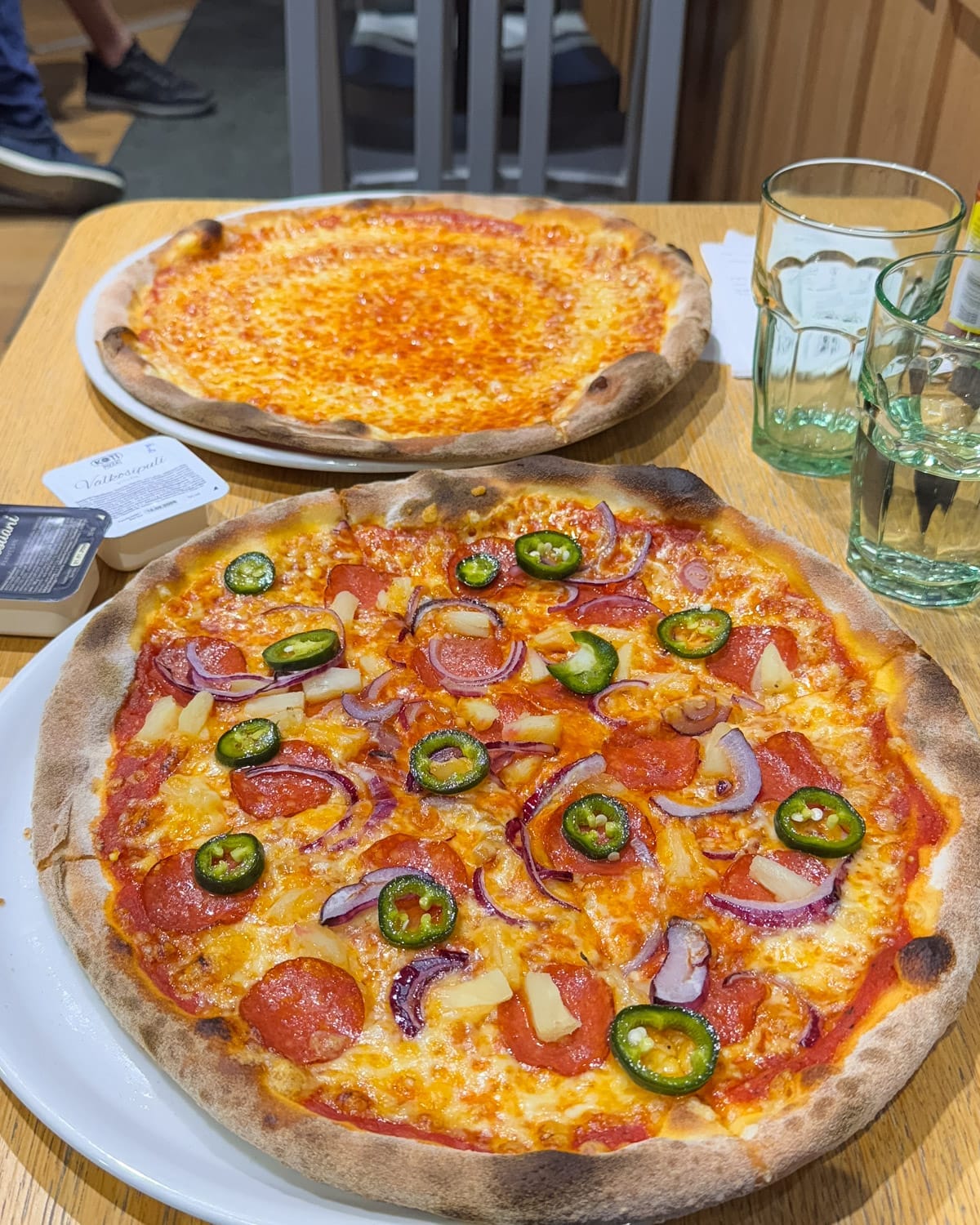 Pepperoni, pineapple, pickled jalapeño, and red onion pizza on a wooden table at Kotipizza in Helsinki.