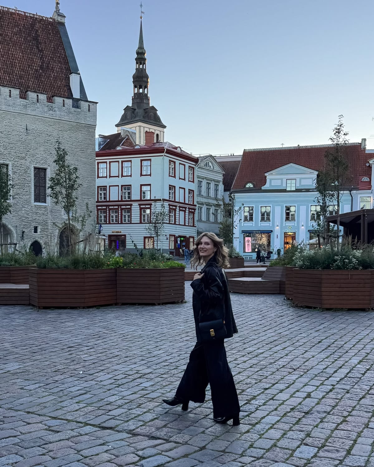 Cec walking across Tallinn’s Town Hall Square at dusk with medieval towers and historic buildings behind her.