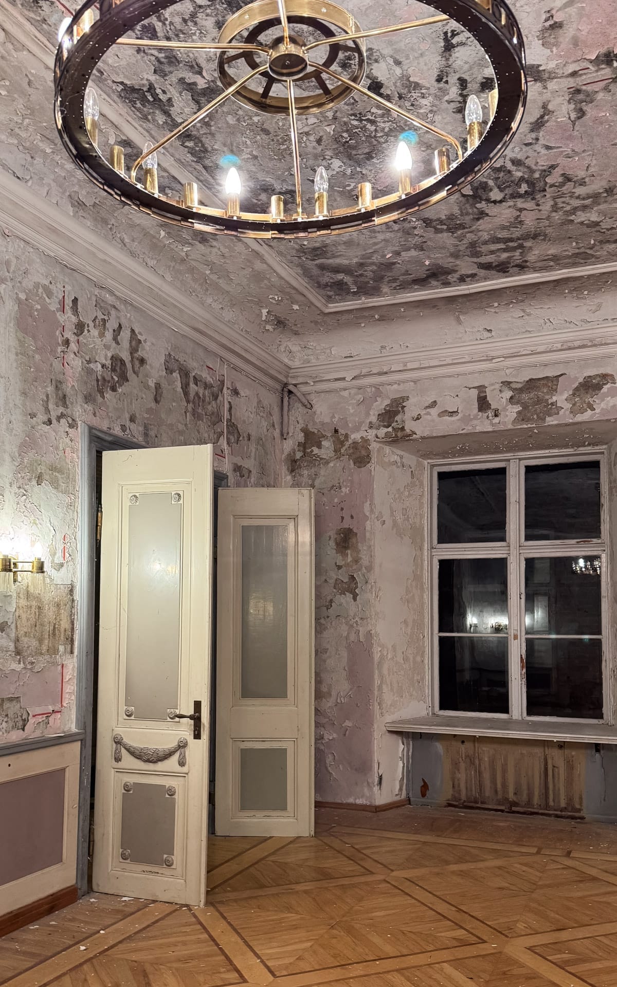 Peeling plaster walls and ornate chandelier inside an abandoned manor room with parquet floors.