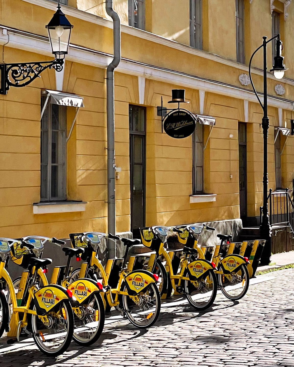 Yellow rental bikes lined up on a cobblestone street in Helsinki, with a mustard-coloured historic building and vintage street lamps in the background.