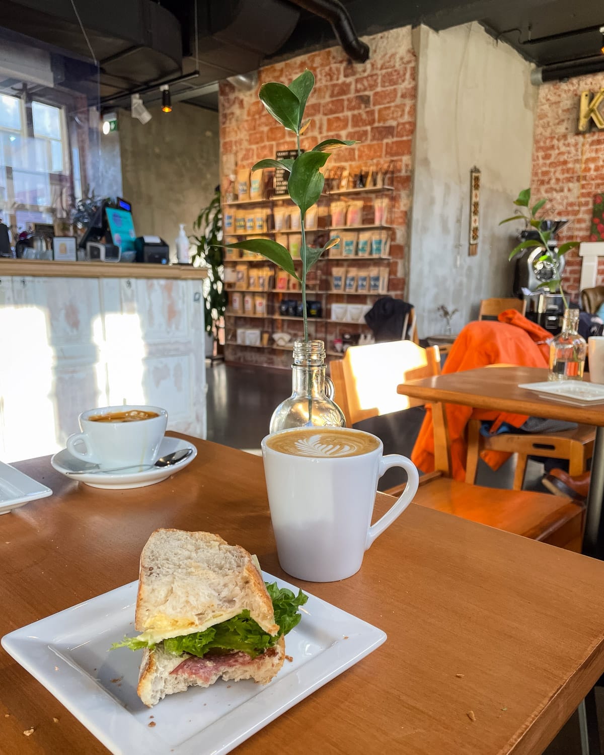 Latte and sandwich on a wooden cafe table inside an industrial-style Helsingin Kahvipaahtimo with brick walls.