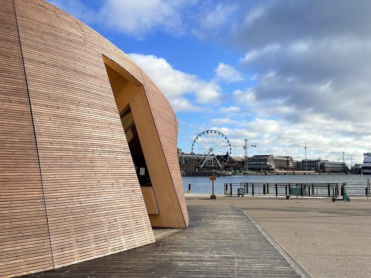 Wooden Helsinki Biennial Pavilion by the waterfront, with harbour views and a ferris wheel in the background.