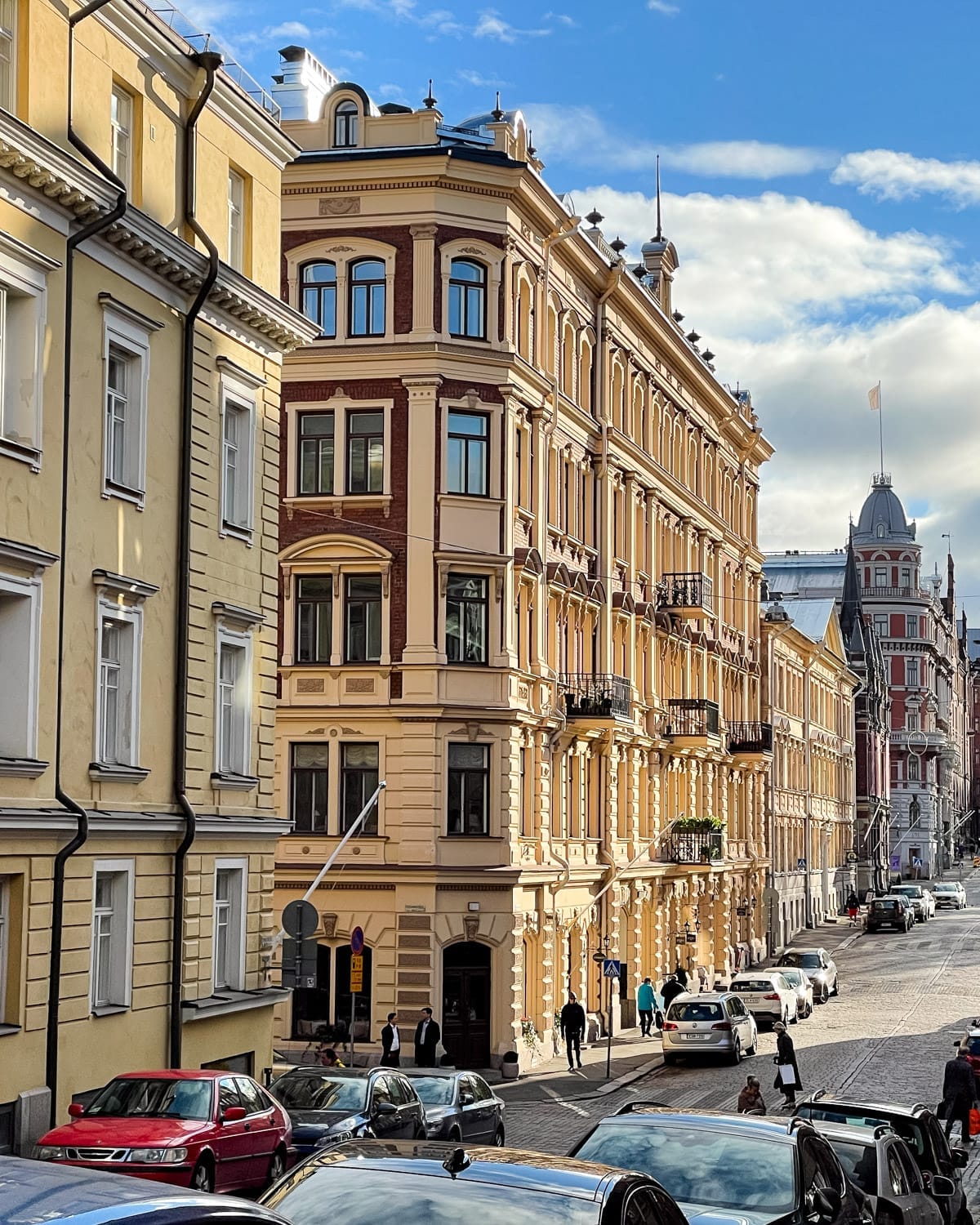 Historic pastel apartment buildings lining a cobblestone street in central Helsinki, with cars parked along the road.