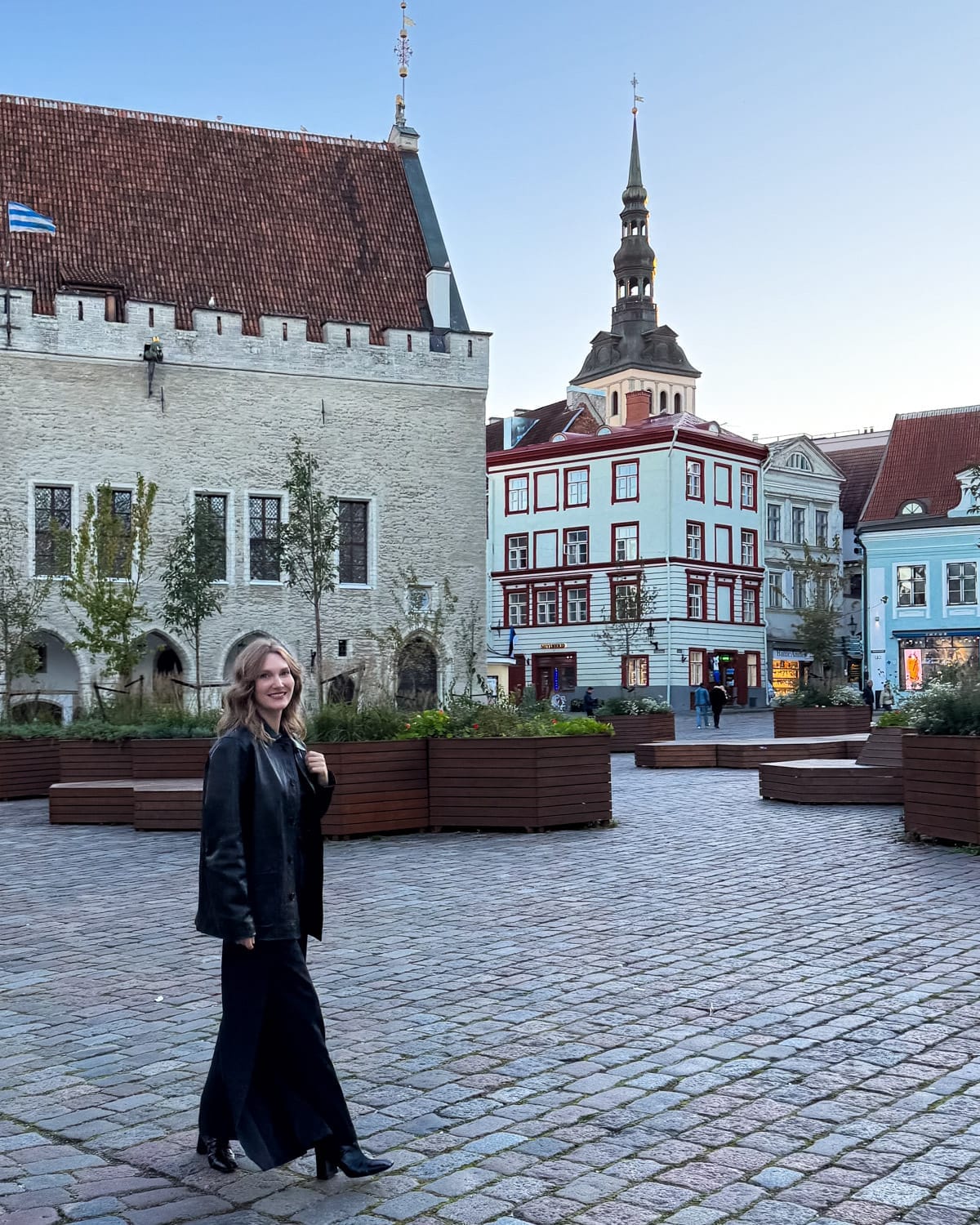 Cec walking across Tallinn’s Town Hall Square at dusk with medieval towers and historic buildings behind her.