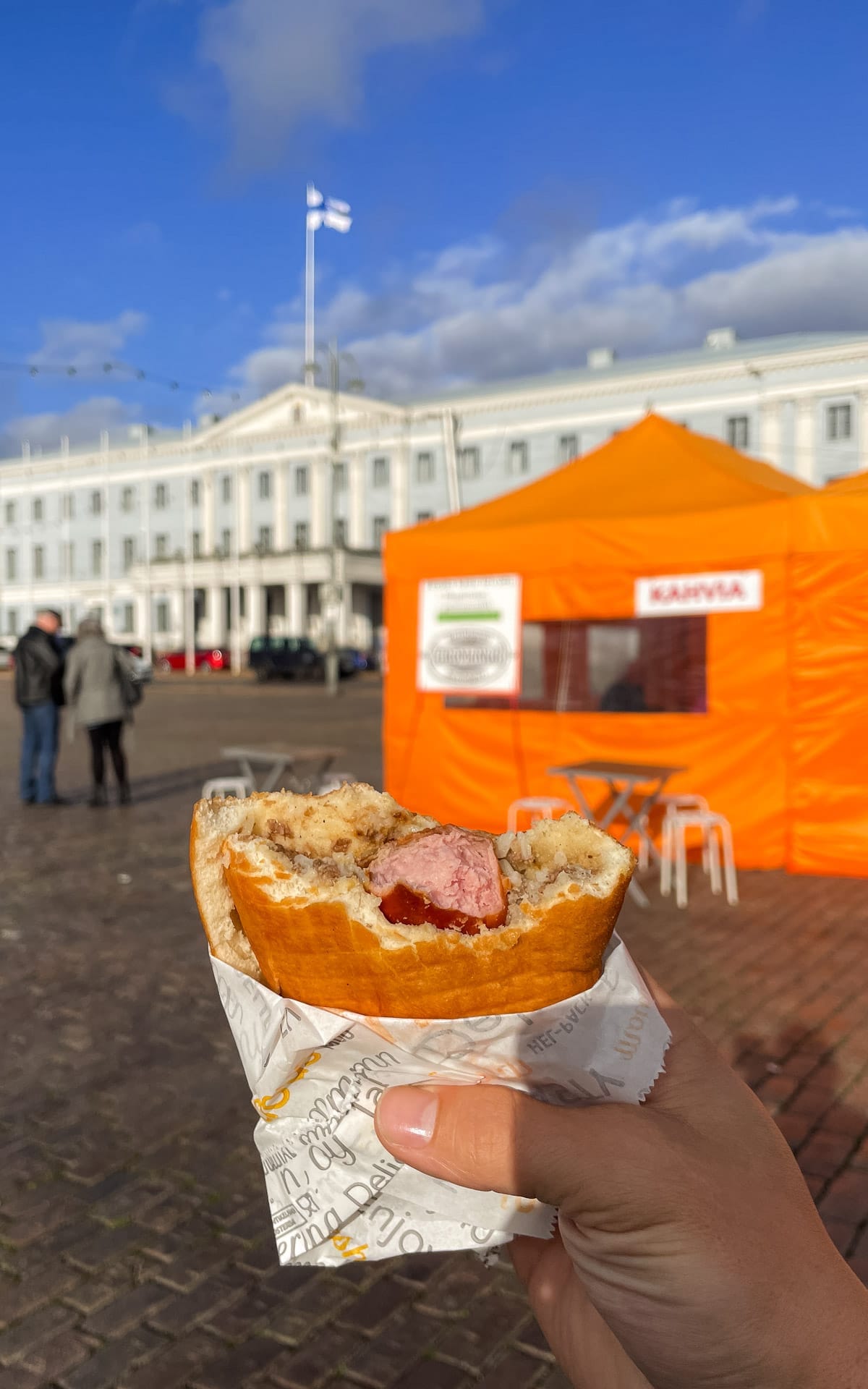 Lihapiirakka meat pie with a hot dog and mustard held in front of the bright orange coffee tent at Helsinki Market Square.