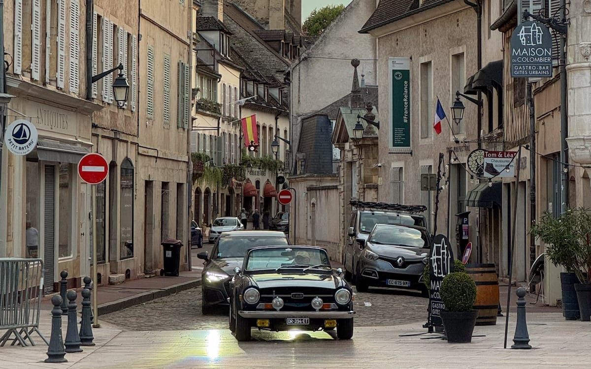 Historic street in Beaune lined with stone buildings and shops, with a vintage car driving on the cobblestones.