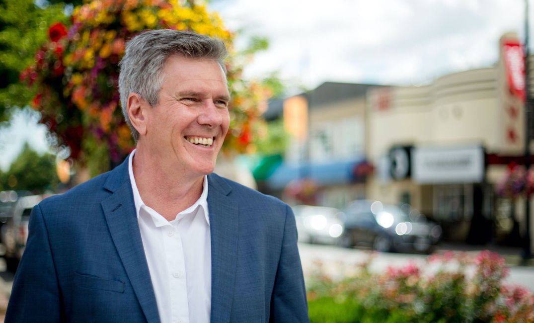 A photo of John Aldag wearing a suit in front of colorful trees