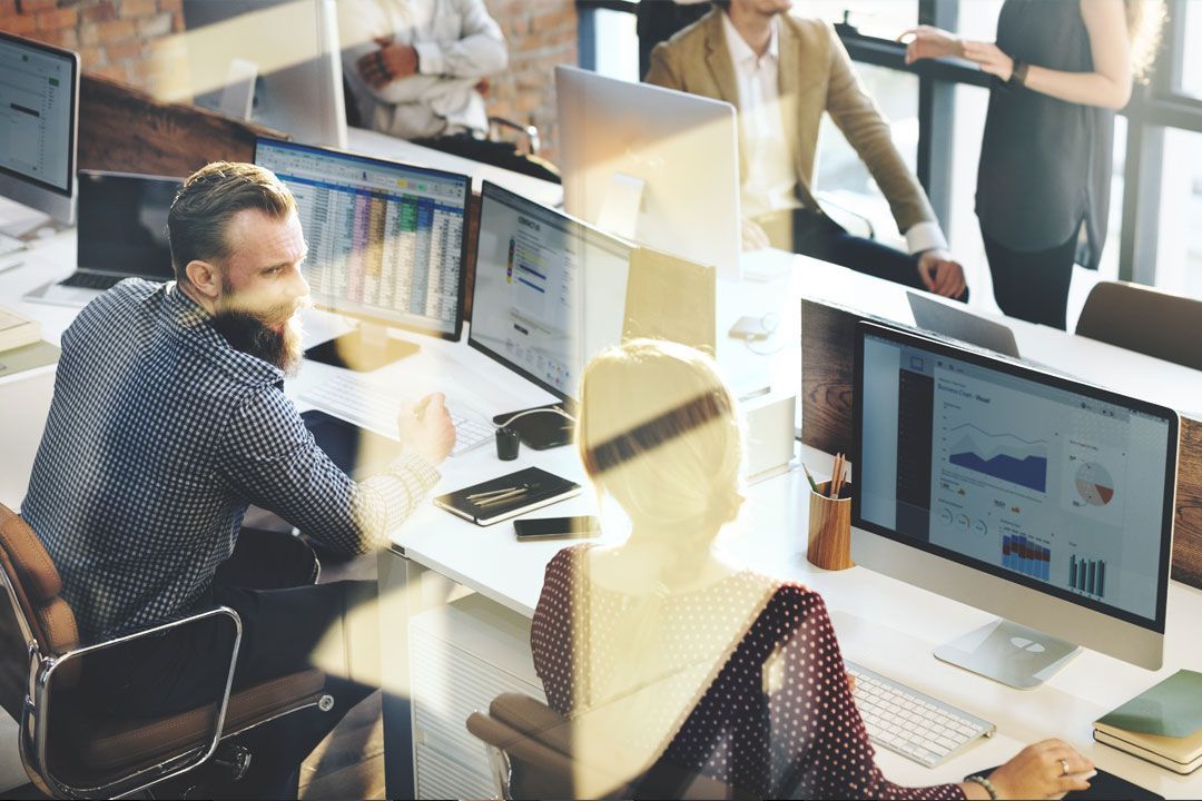 Teammates in an open-plan office chatting in front of their computers. 
