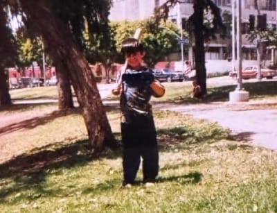 A young boy, Steven Renderos, holds a baseball bat and glove in a city park.