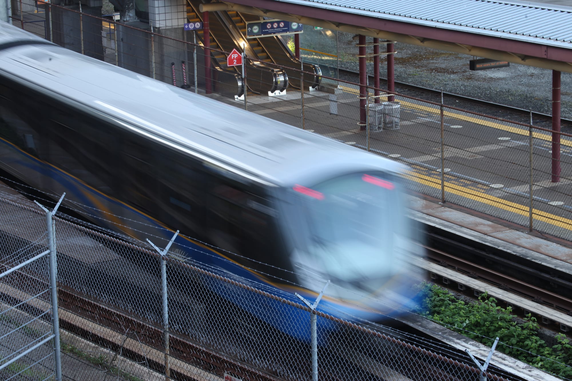 A SkyTrain speeds past blurred at an empty platform surrounded by barbed wire fences