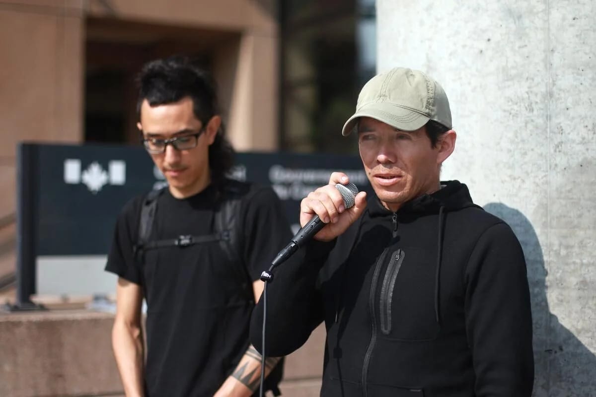 A man in a baseball cap speaks into a microphone, with another person standing beside him and a Canadian government office in background