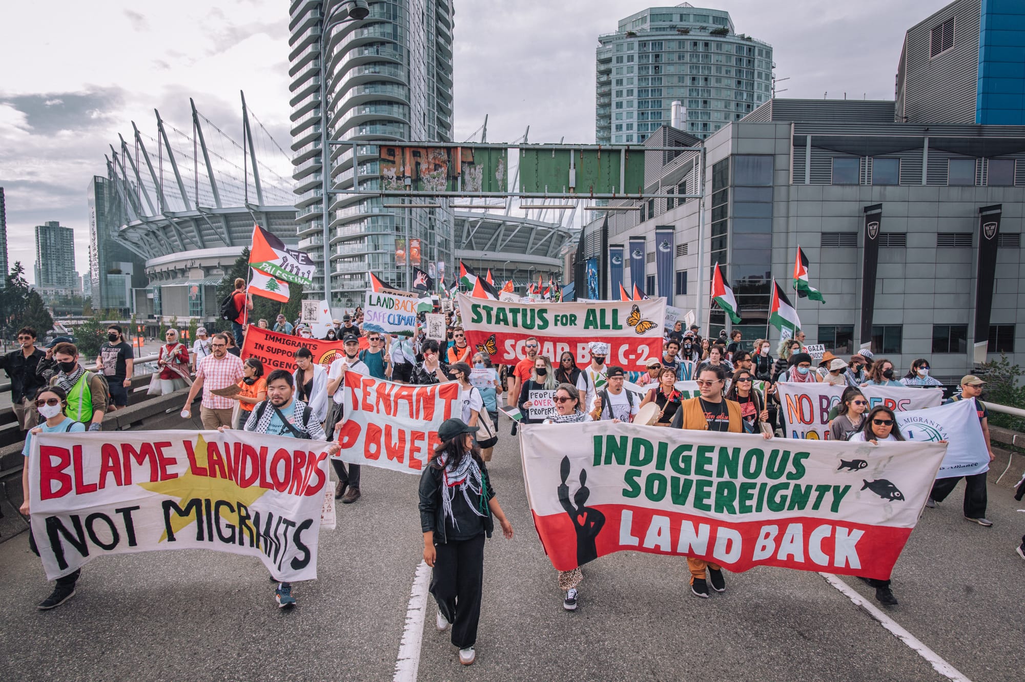 Protesters walk on a roadway with a city behind, carrying banners saying "Indigenous sovereignty, land back," and "Status for all"