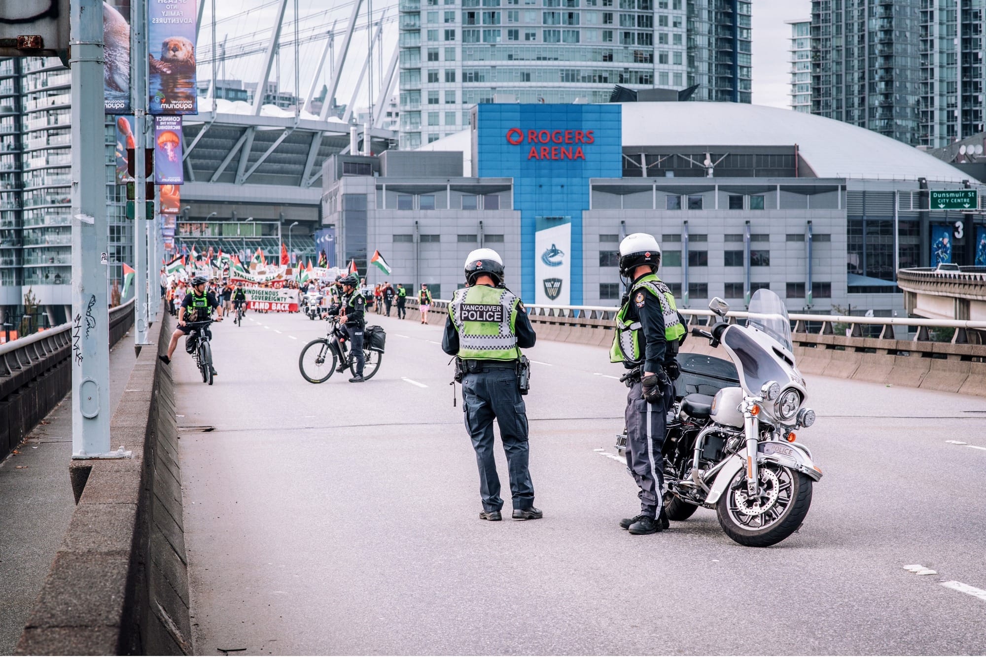 Four police officers stand by a police motorbike and police bicycles on a bridge, as protesters approach with banners and flags in the distance