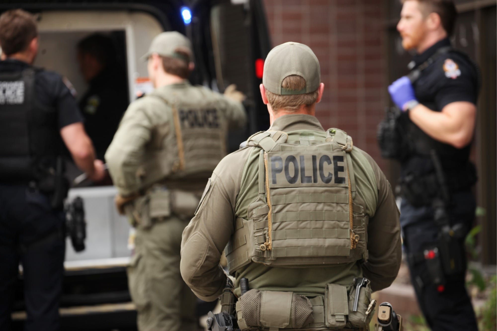 Four police officers, two in olive-green tactical uniforms, and two in standard uniforms, stand behind a police van.