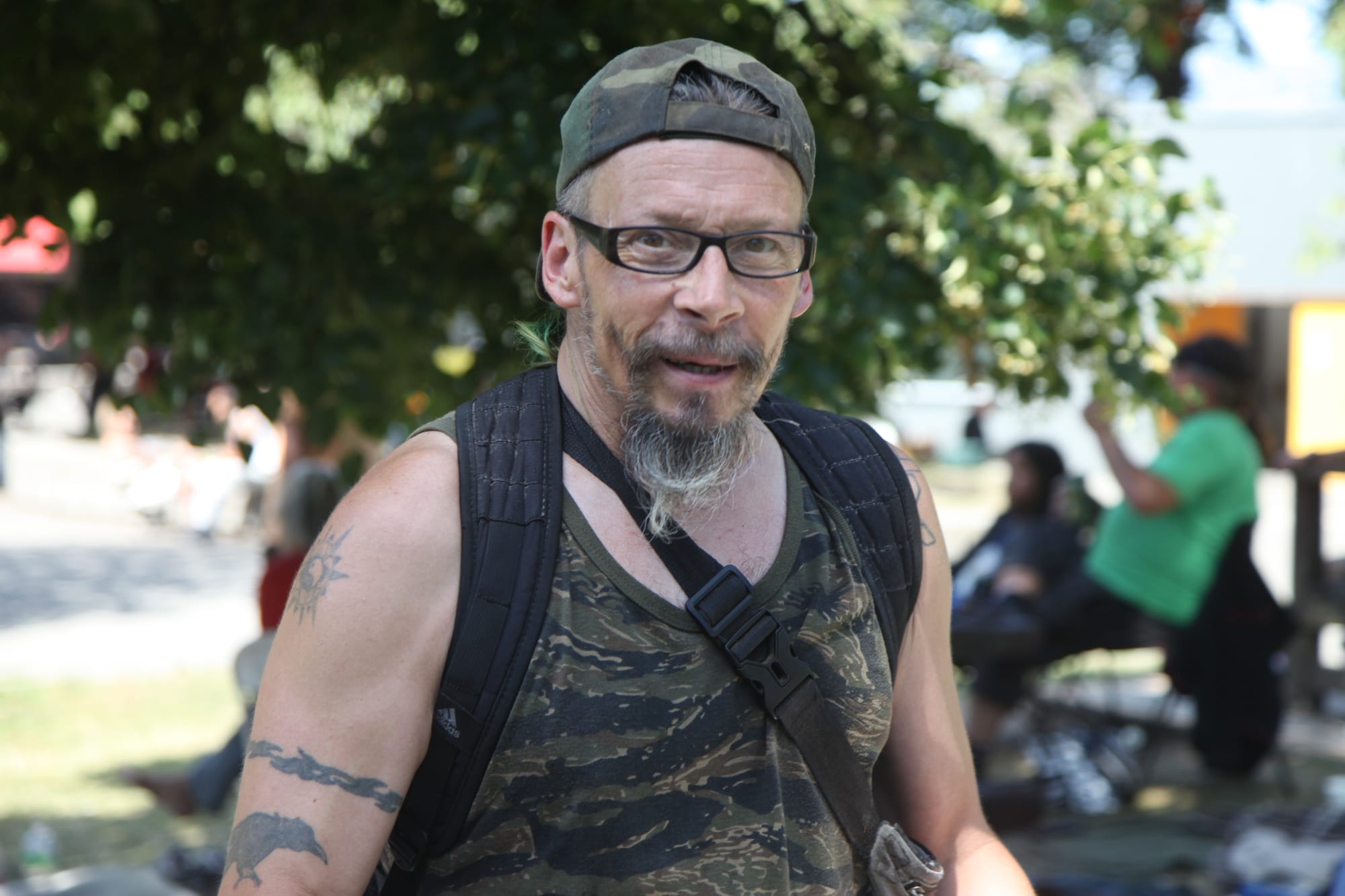Harley in a park wearing glasses, a greying goatee, a camo hat and tank top, and a backpack and bag. 