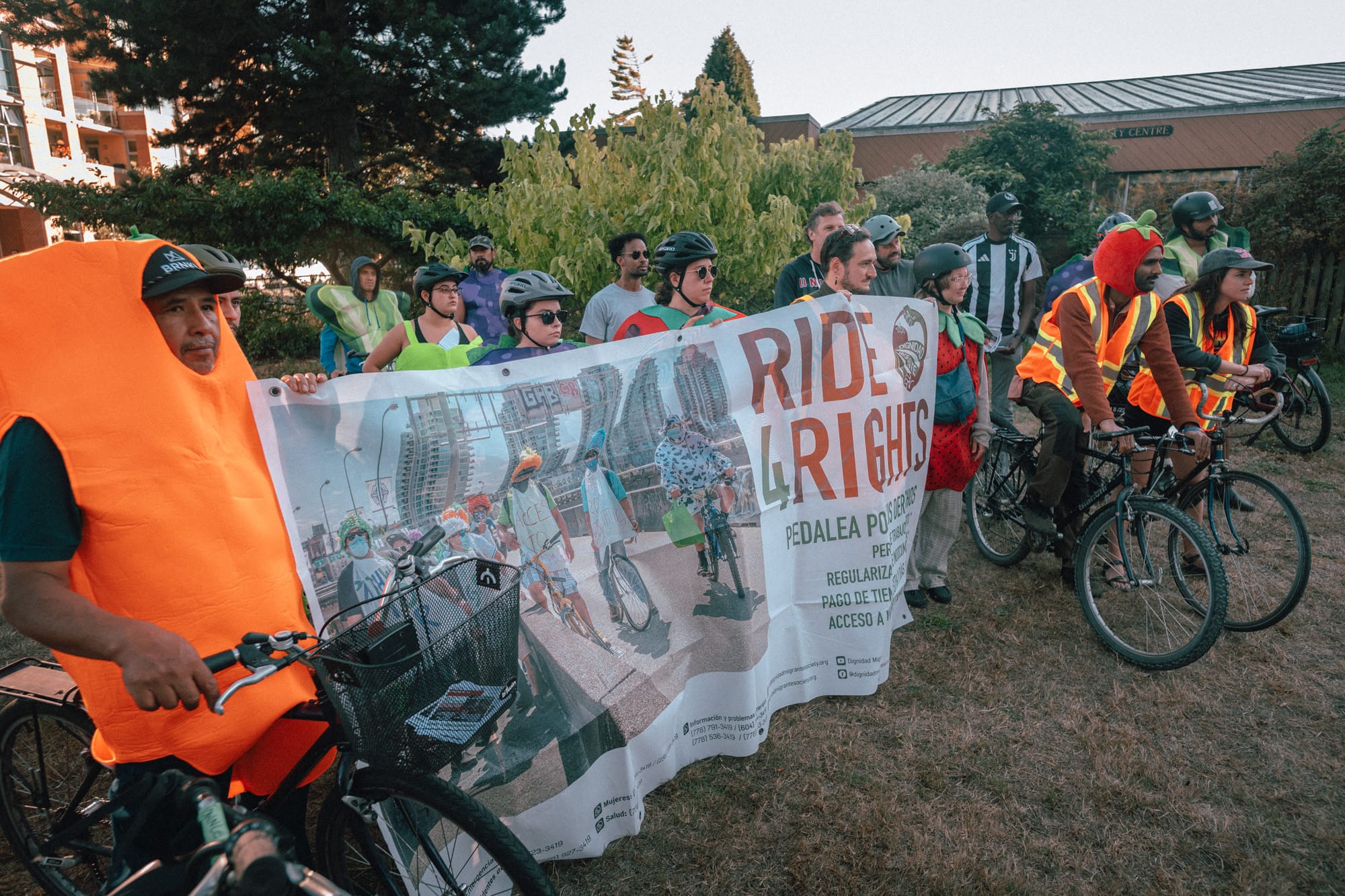Riders in hi-vis vests and fruit and veggie costumes pose on bikes with the Ride4Rights banner in James Bay