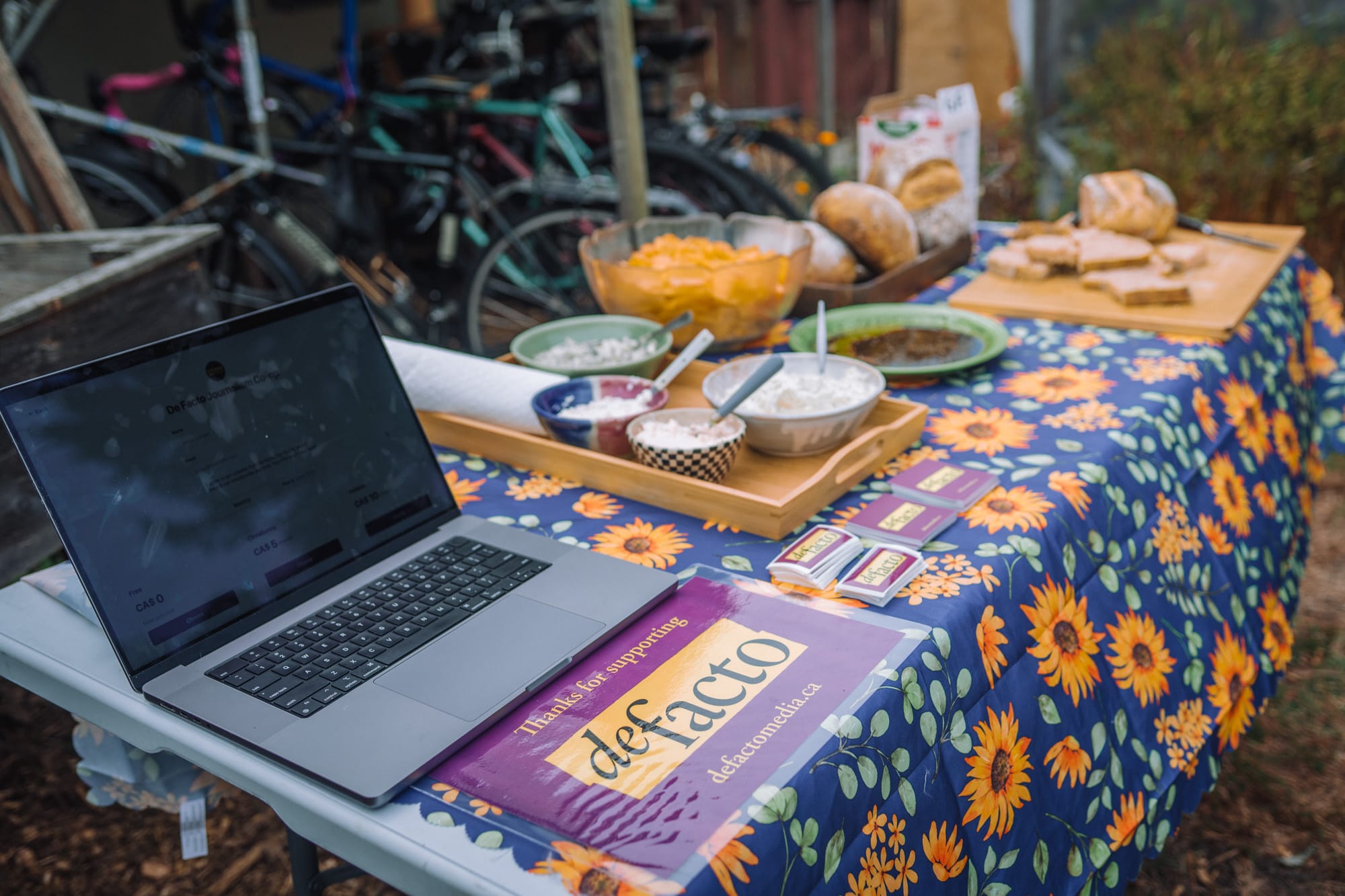 A laptop, purple sign, and food on a blue sunflower tablecloth covering a backyard table