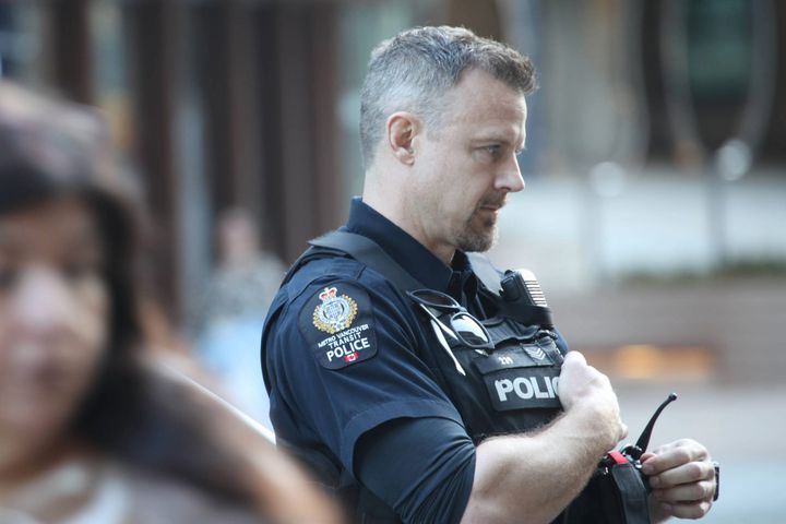 A Metro Vancouver Transit Police officer stands outside looking away as a woman walks past