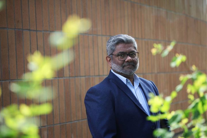 A lawyer wears a blue suit behind some leaves with a brick wall in the background