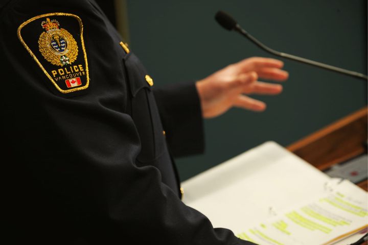 A Vancouver Police officer's shoulder and raised hand are seen, above a highlighted document, as the officer speaks at a lectern during a press conference 