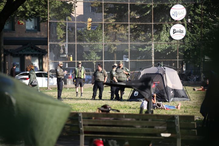 Park officers gather beside a tent near a baseball fence, seen from across the park