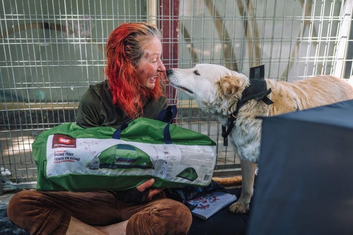 A woman with red dyed hair touches noses with a golden retriever type dog as she holds a tent