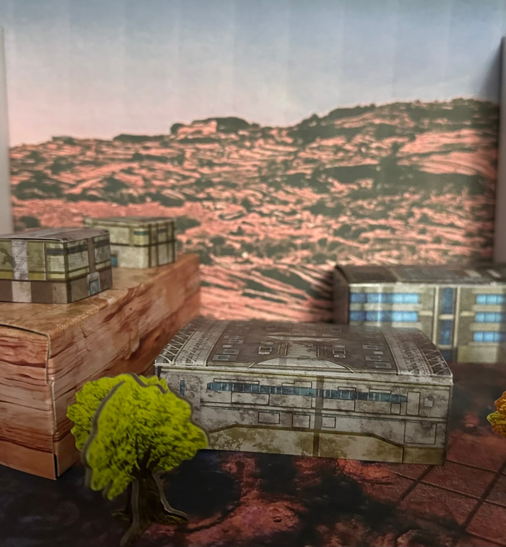 Card terrain of a several buildings, a hill and a tree on a red desert floor with a rocky background image.
