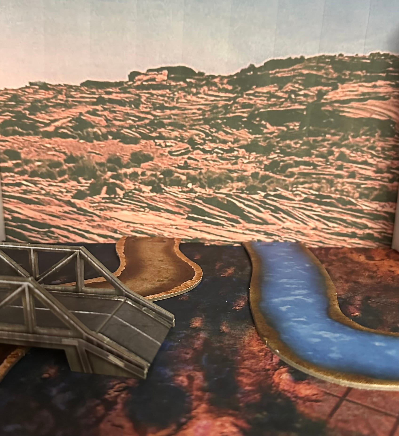 Card terrain of a bridge, gully and river on a red desert floor with a rocky background image.