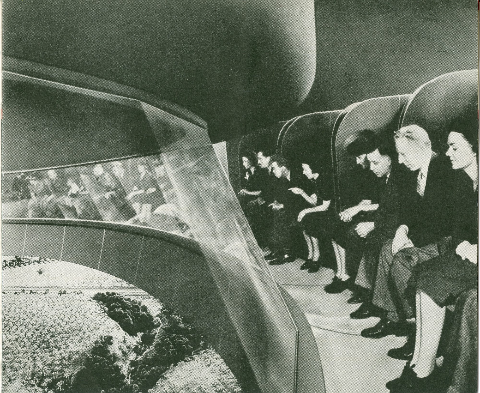 Visitors in formal 1930s attire seated in the curved, modernist viewing area of the Futurama pavilion at the 1939 World's Fair, looking down at an expansive miniature landscape model below. The streamlined architecture features flowing organic curves, and guests appear captivated by the detailed diorama displaying a vision of future America.