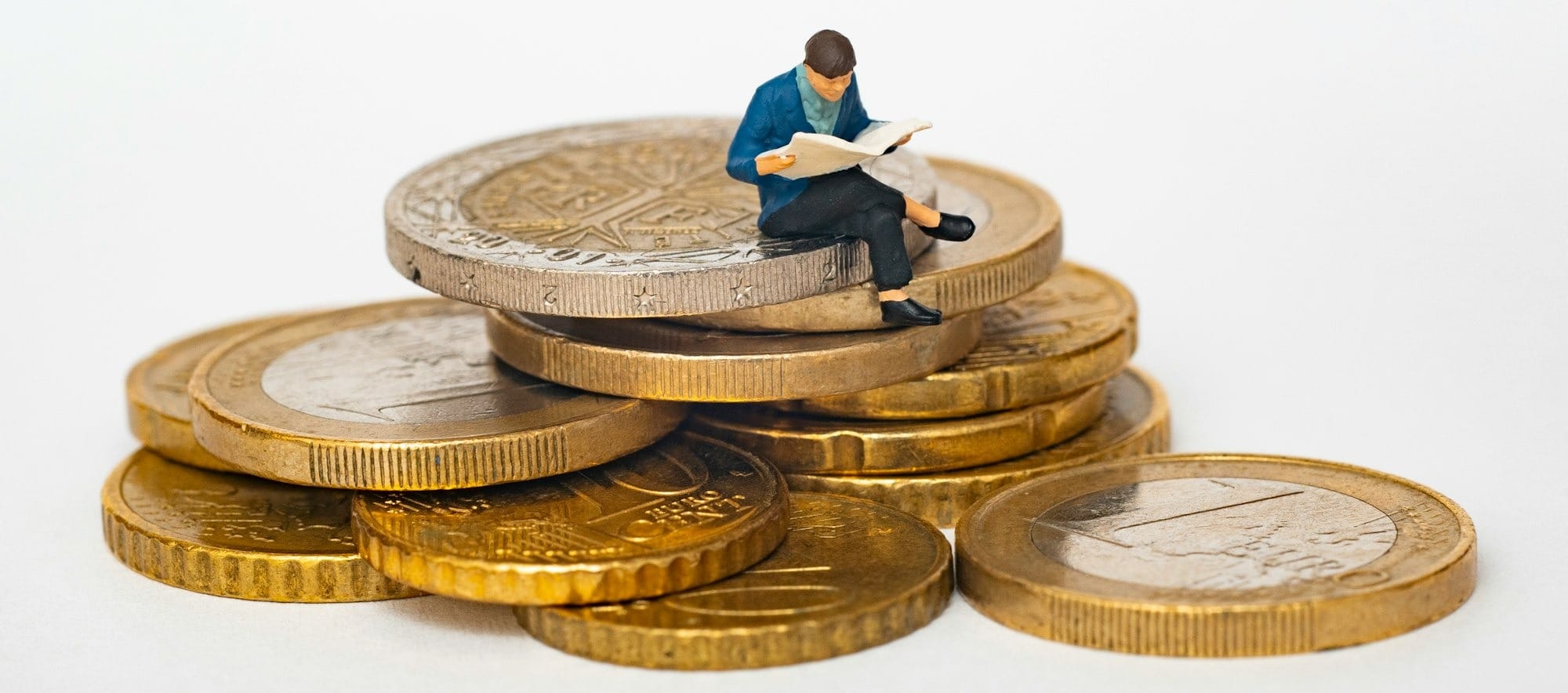 Miniature person sitting on stack of coins reading newspaper
