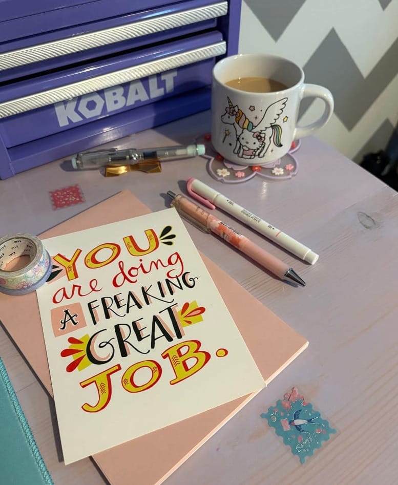Pens, washi tape, and card that reads "You are doing a freaking great job". In the background, a mug with Hello Kitty and unicorn filled with coffee